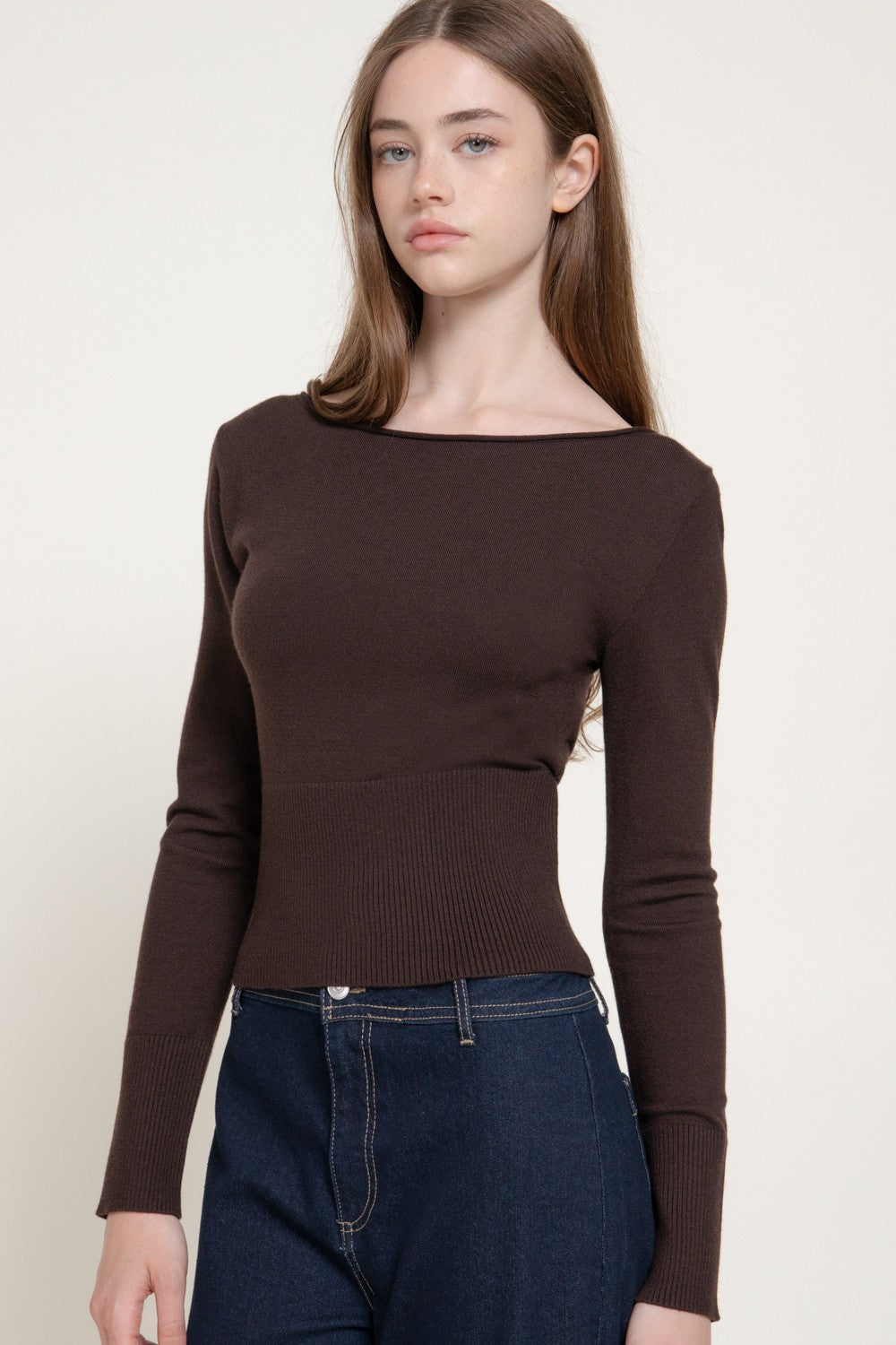 A woman modeling a dark brown ribbed knit long-sleeve top with a wide boat neckline, paired with dark blue jeans, against a plain off-white background.