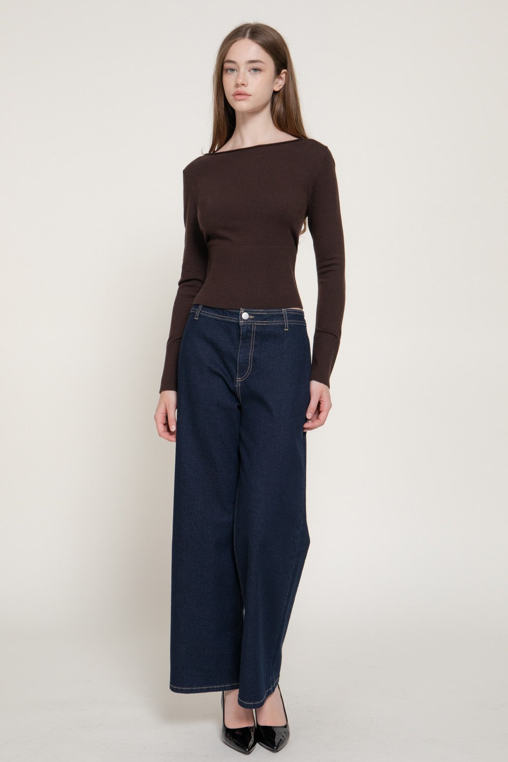 A woman modeling a dark brown off-the-shoulder long-sleeve top paired with dark blue wide-leg jeans and black pointed heels, standing against a plain light gray studio background.