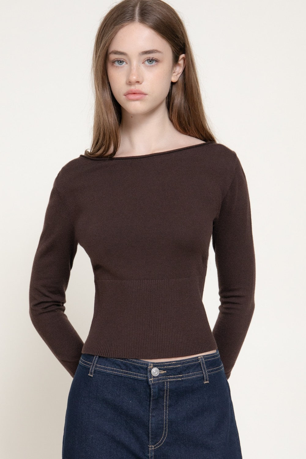 A woman modeling a dark brown ribbed knit long-sleeve top with a wide boat neckline, paired with dark blue jeans, against a plain off-white background.
