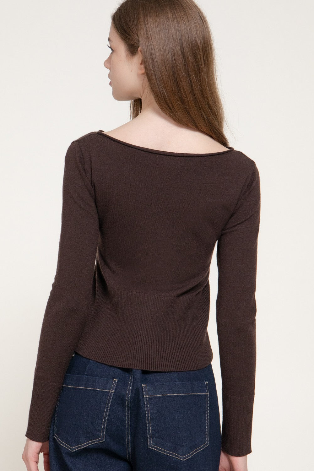 A woman shown from the back wearing a dark brown long-sleeve knit top with a wide boat neckline and dark blue jeans, against a plain light background.