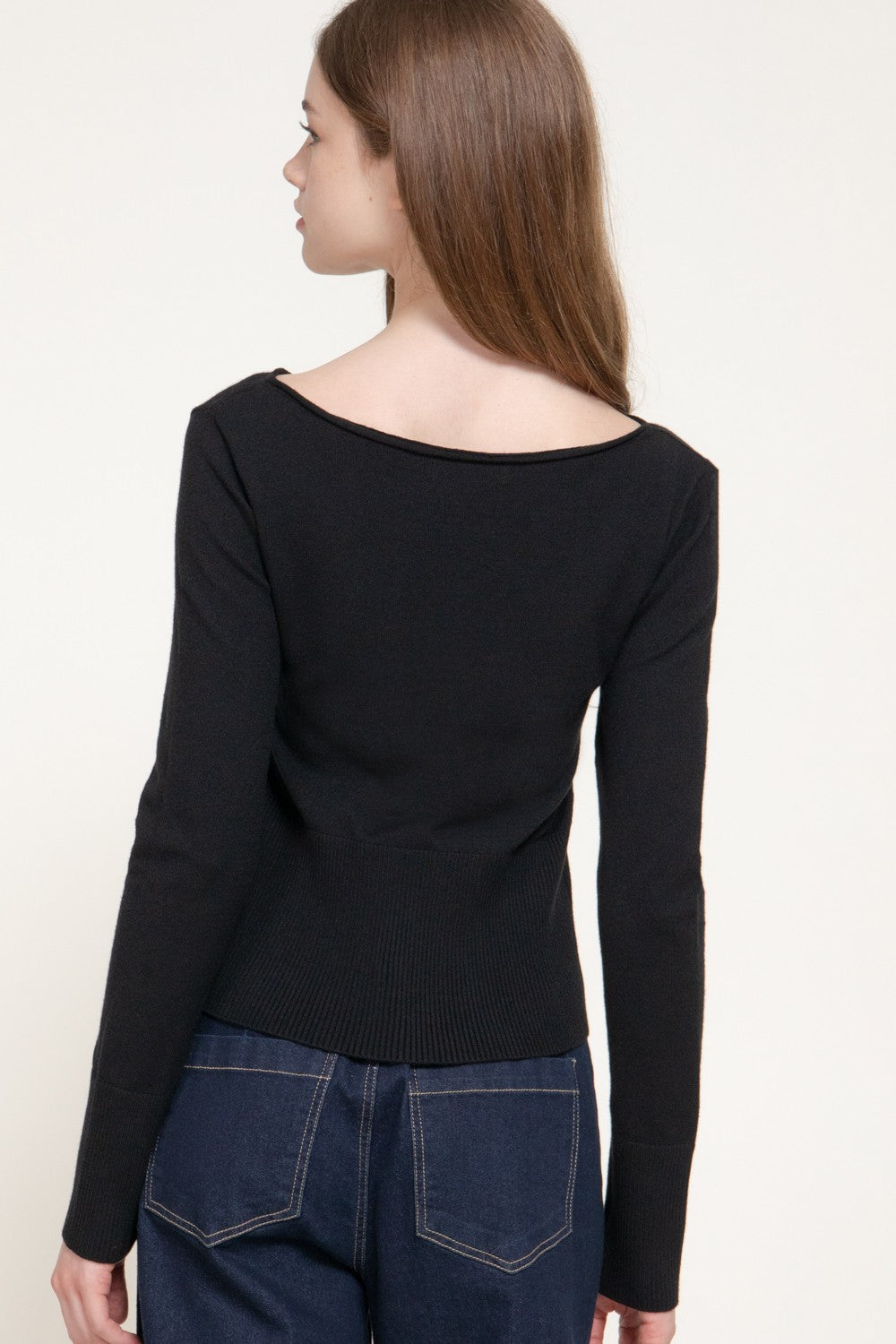 A woman shown from the back wearing a black long-sleeve knit top with a wide boat neckline and dark blue jeans, against a plain light background.