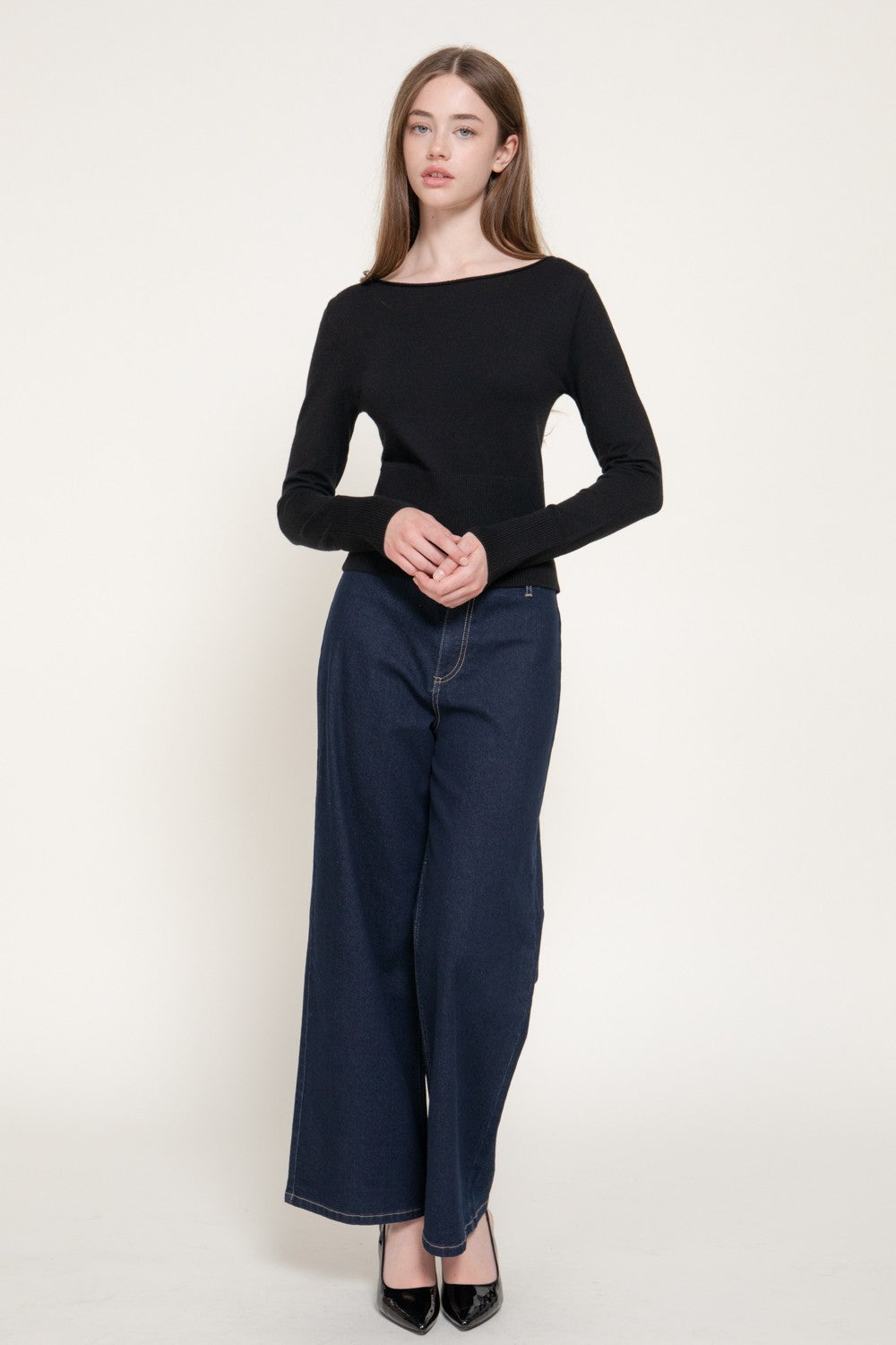 A woman modeling a black long-sleeve boat-neck top paired with dark blue wide-leg jeans and black pointed heels, standing against a plain off-white studio background.