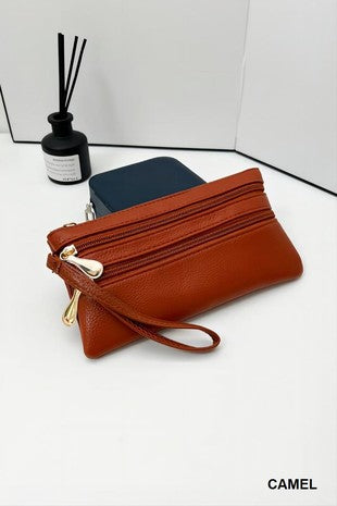 A camel brown leather zip-around wristlet pouch with gold-tone hardware, displayed on a white surface next to a black reed diffuser and a dark blue pouch. The word 'CAMEL' is printed in the bottom right corner.