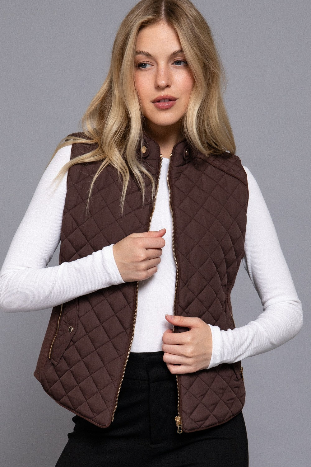 A woman wearing a brown diamond-quilted zip-front vest over a white long-sleeve top and black pants, photographed against a plain gray studio background.