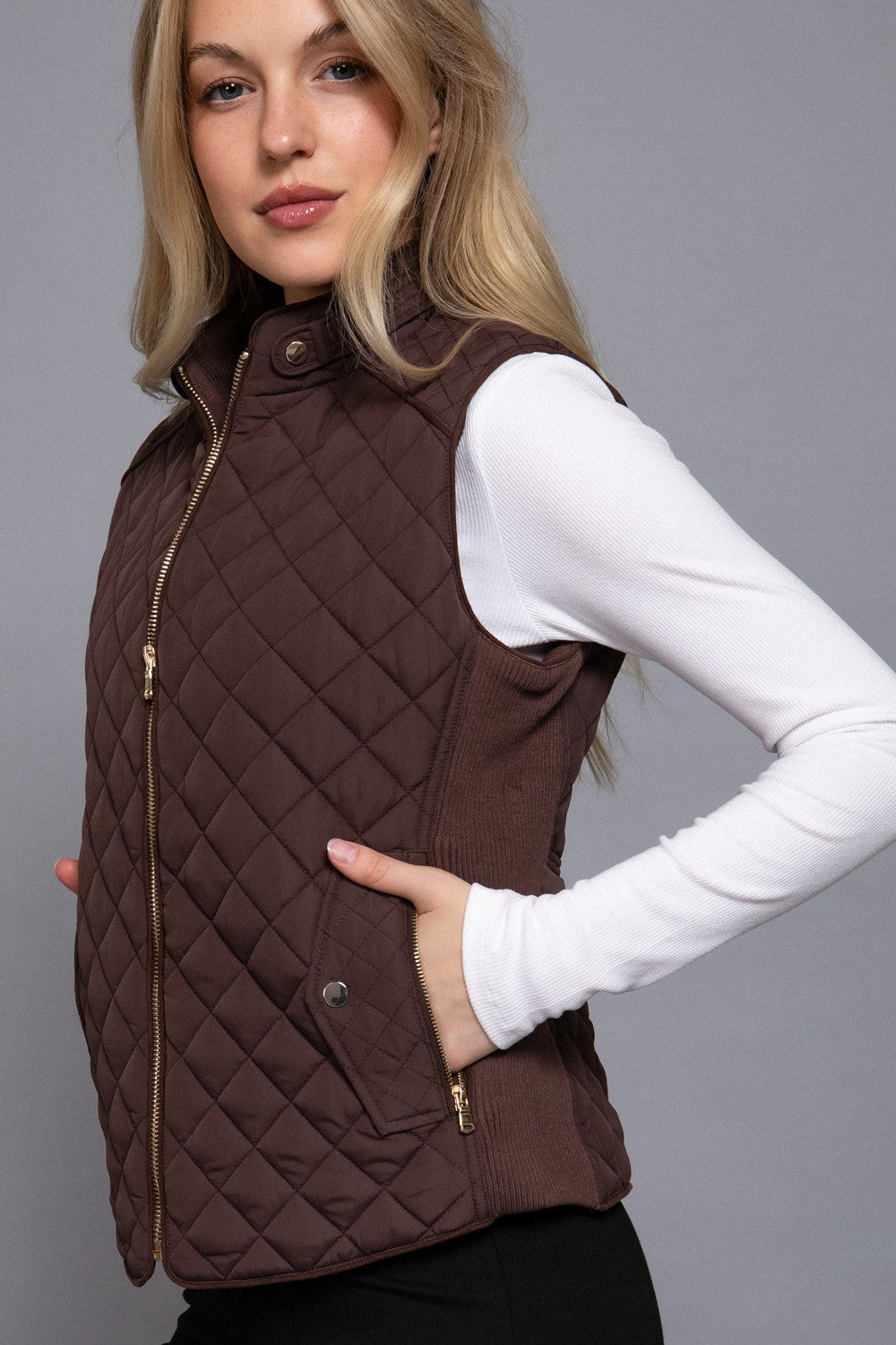 A woman wearing a brown diamond-quilted zip-front vest over a white long-sleeve top, photographed against a plain gray studio background.