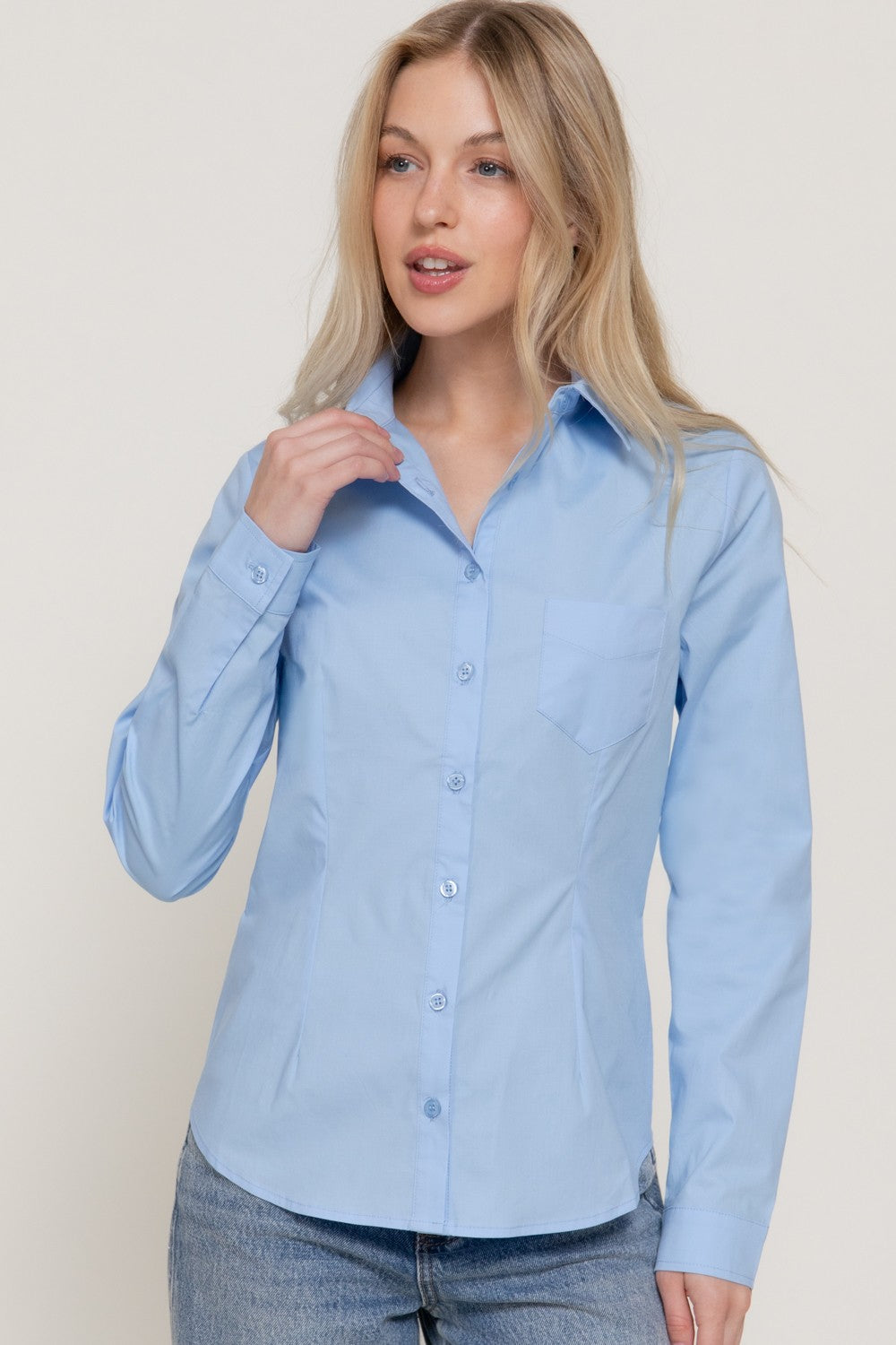 A woman wearing a light blue long-sleeve button-up shirt and blue jeans, standing against a plain light beige studio background.
