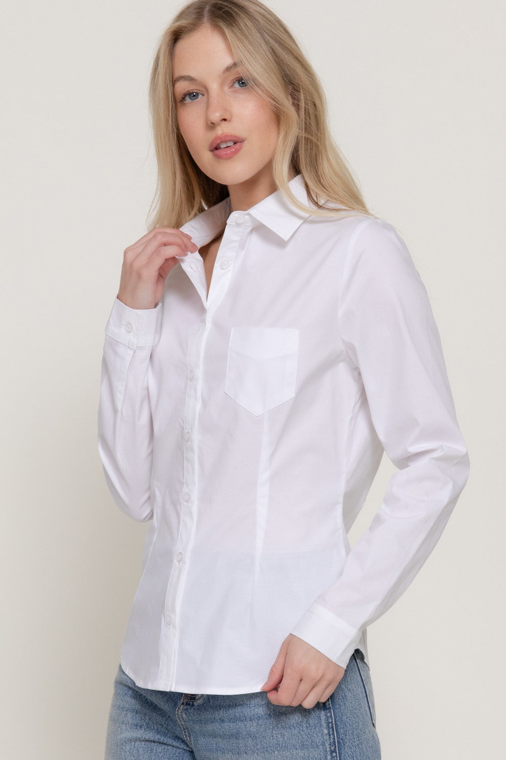 A woman wearing a white long-sleeve button-up shirt with a chest pocket, paired with blue jeans, photographed against a plain light beige studio background.