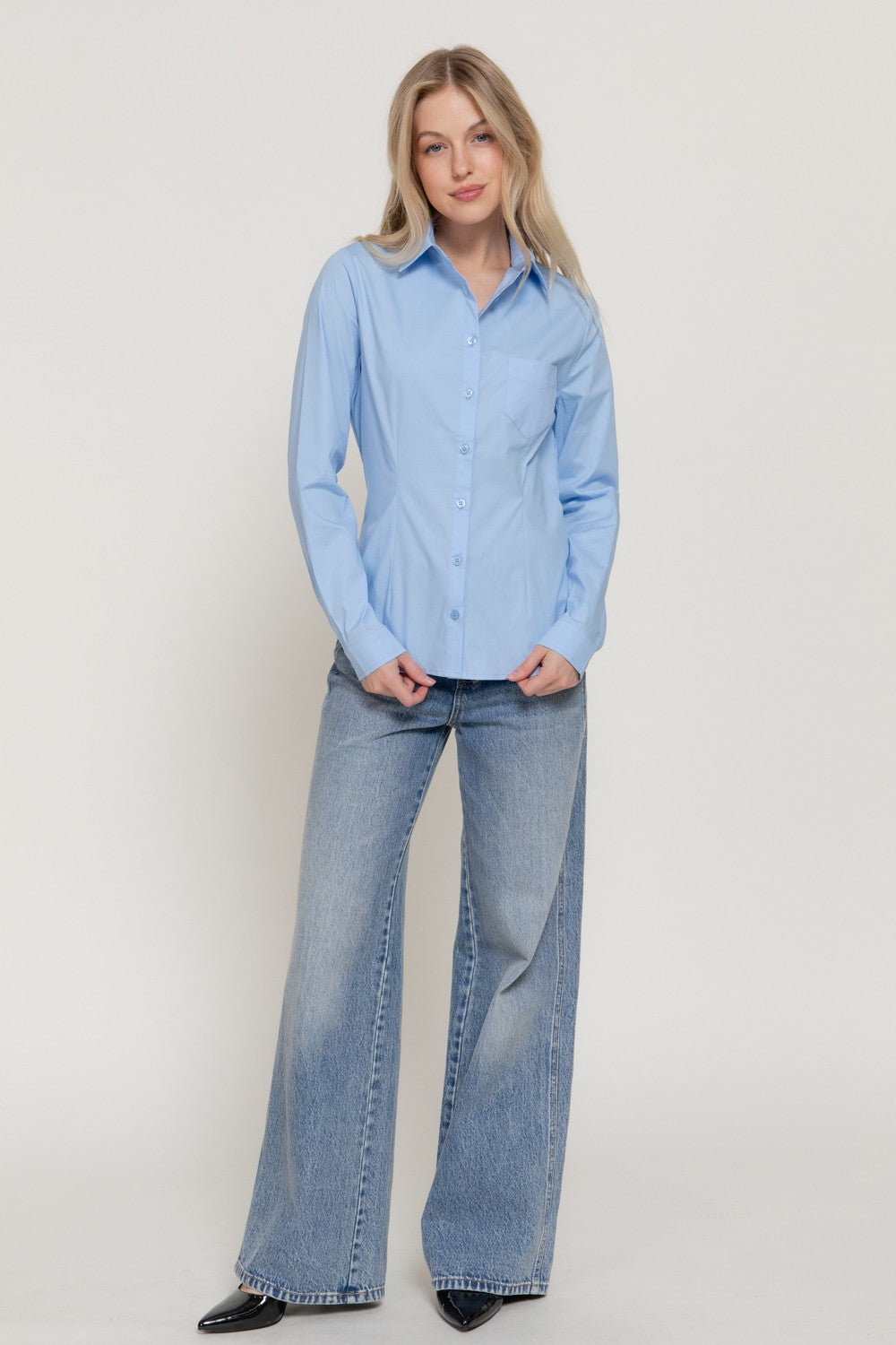 A woman modeling a light blue long-sleeve button-up shirt with wide-leg light-wash blue jeans, standing against a plain light gray studio background.