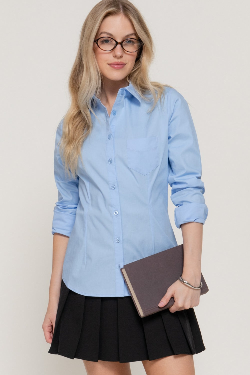 A woman with blonde hair and glasses wearing a light blue long-sleeve button-down shirt and a black pleated skirt, holding a dark brown notebook against a light beige background.