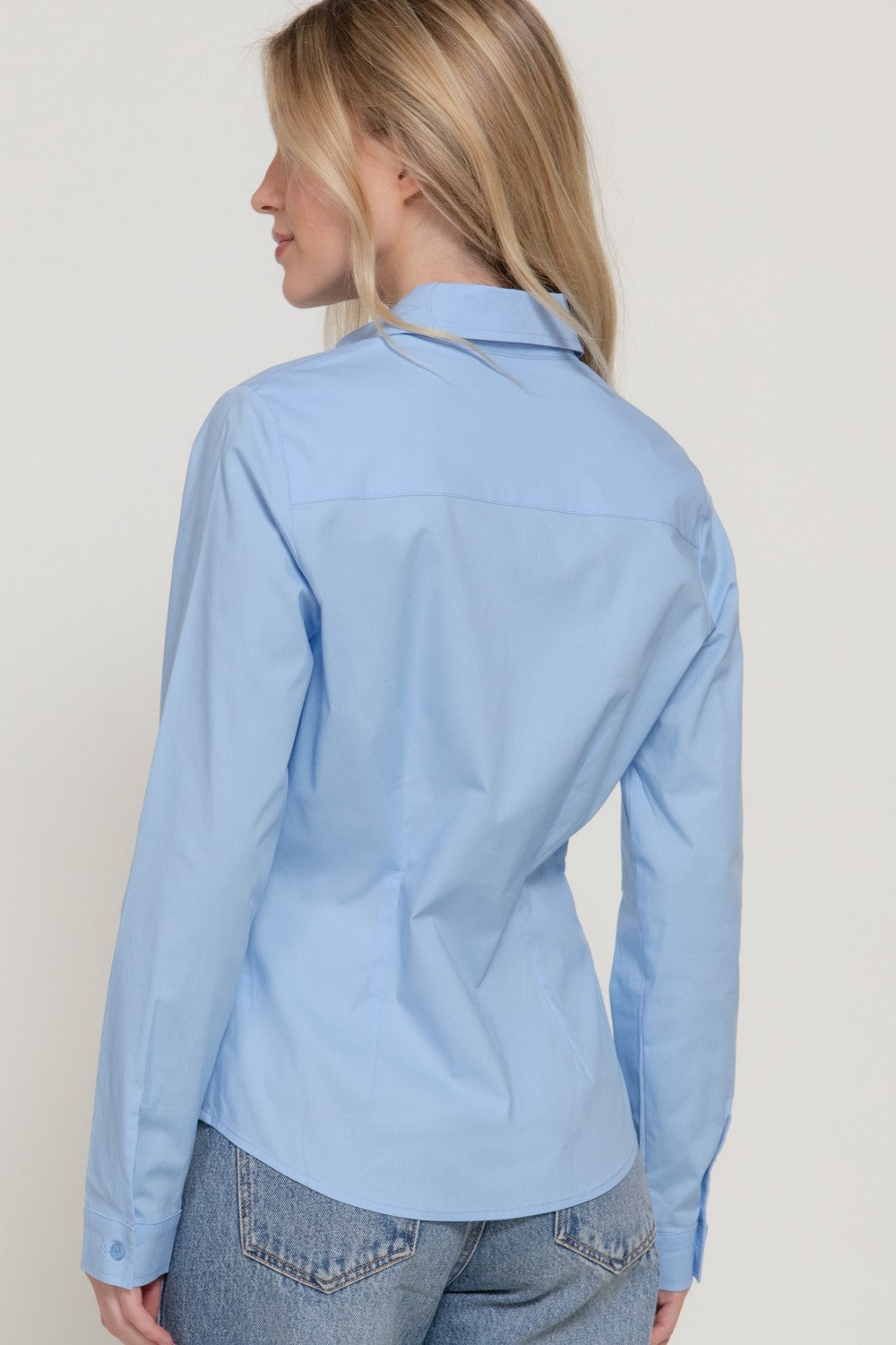 A woman shown from the back wearing a light blue long-sleeve button-up shirt and light-wash blue jeans against a plain light background.