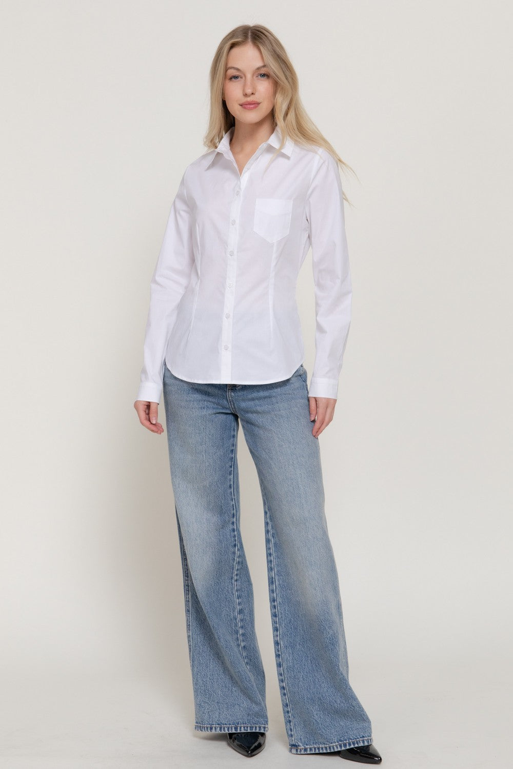 A woman modeling a white long-sleeve button-up shirt tucked into light-wash blue wide-leg jeans, standing against a plain light beige studio background.