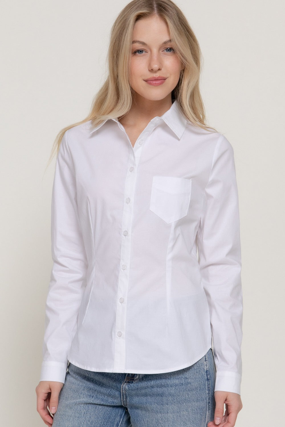 A woman wearing a white long-sleeve button-up shirt with a chest pocket, paired with blue jeans, photographed against a plain light beige studio background.