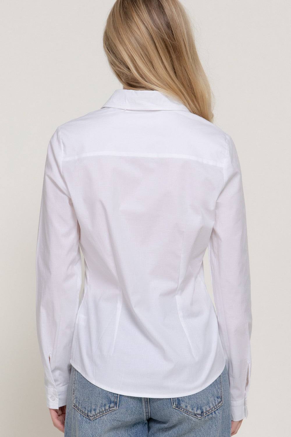 A woman viewed from the back wearing a white long-sleeve button-up shirt and light blue jeans against a plain light beige background.