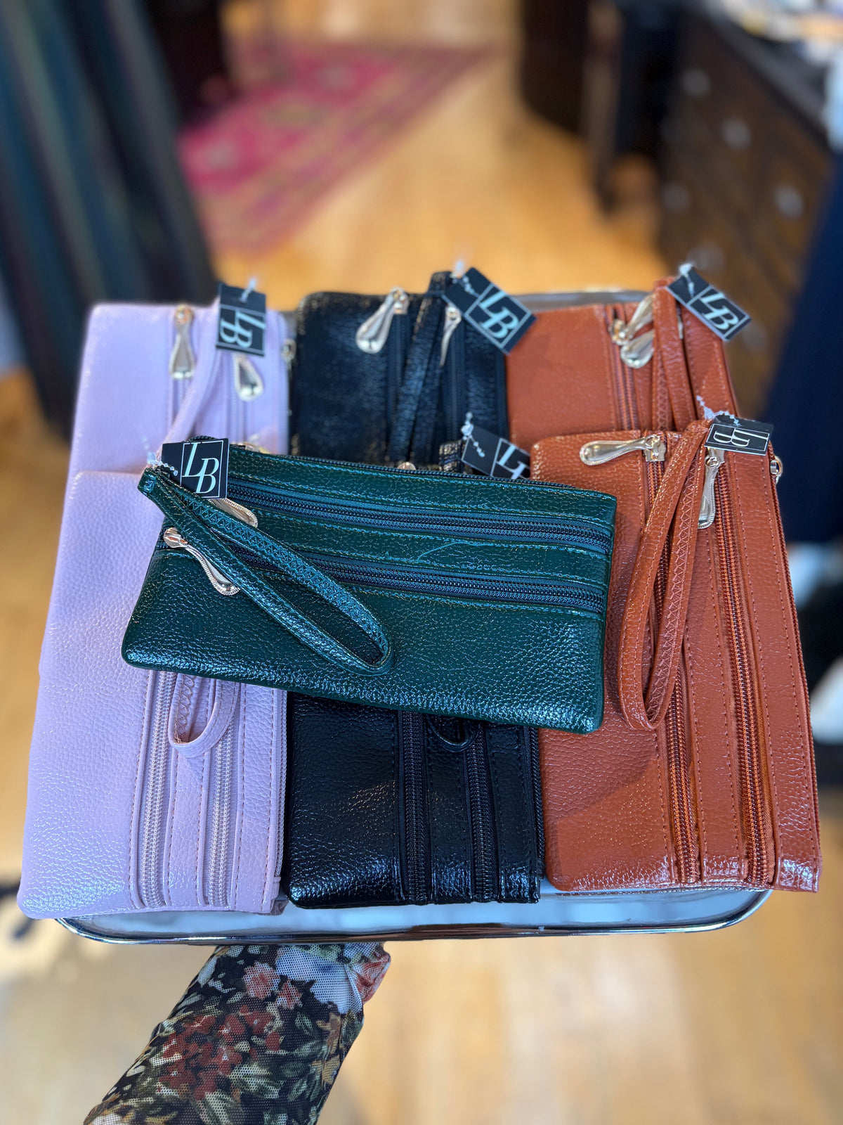 A person holds a tray displaying multiple faux leather zip-around card holders and wristlet pouches in light purple, black, and rust brown, with a dark green textured pouch placed on top.