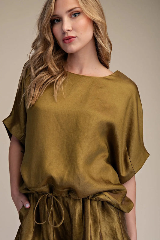 A woman wearing a loose, short-sleeved olive green top with a drawstring waist, photographed against a plain beige studio background.