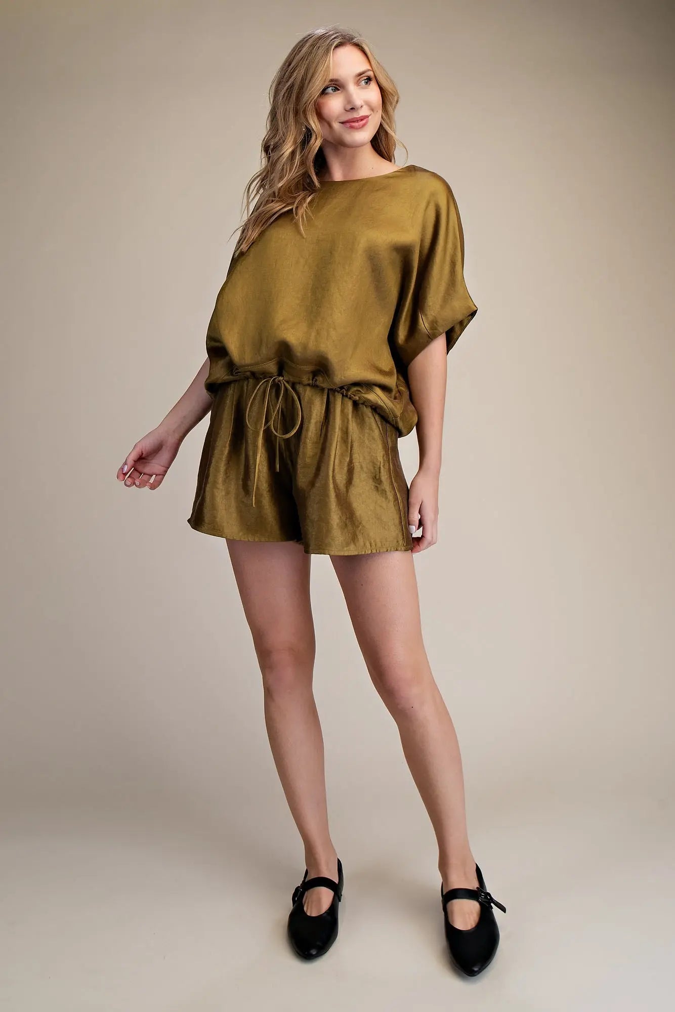 A woman modeling a short, loose-fitting olive green romper or dress with short wide sleeves and a drawstring waist, paired with black Mary Jane shoes, against a plain beige studio background.