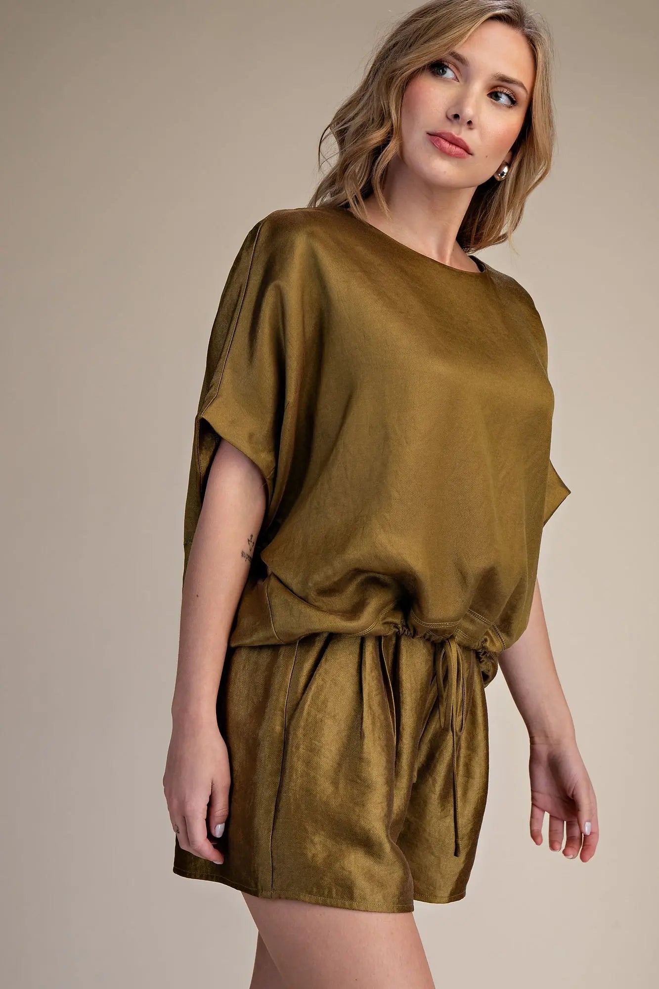 A woman modeling a matching olive green short-sleeve top and shorts set with a drawstring waist, photographed against a plain beige studio background.