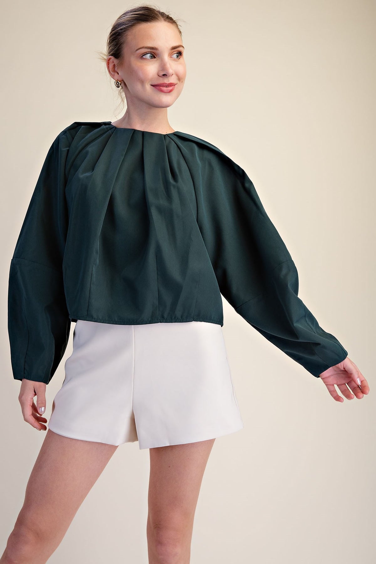 A woman modeling a dark green long-sleeve blouse with a gathered, balloon-like neckline and white shorts against a plain beige studio background.