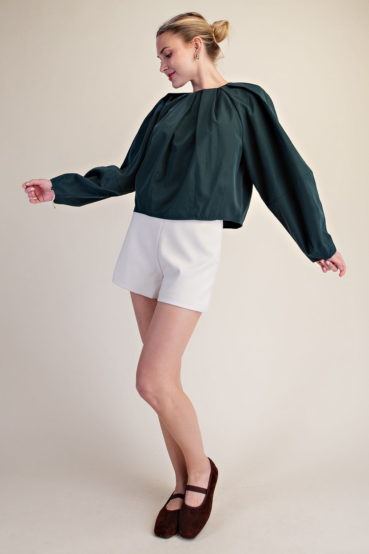 A woman modeling a dark green long-sleeve blouse with dramatic puffed shoulders and white shorts, paired with dark brown flat shoes, against a solid light beige background.