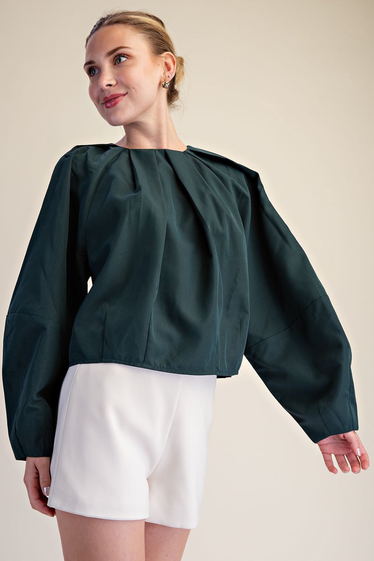 A woman modeling a dark green long-sleeve blouse with a gathered, balloon-like sleeve and pleated neckline, paired with white shorts, against a plain beige studio background.