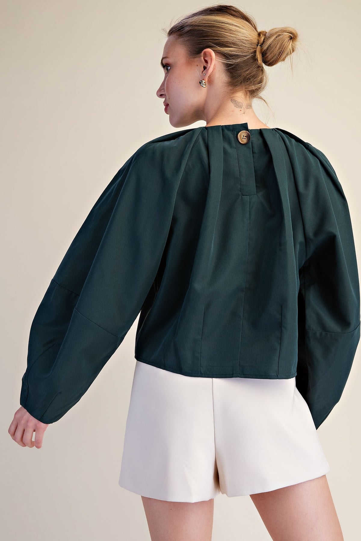 A woman shown from the back wearing a dark green cropped blouse with dramatic balloon sleeves and a single gold button at the neckline, paired with white shorts against a plain beige studio background.