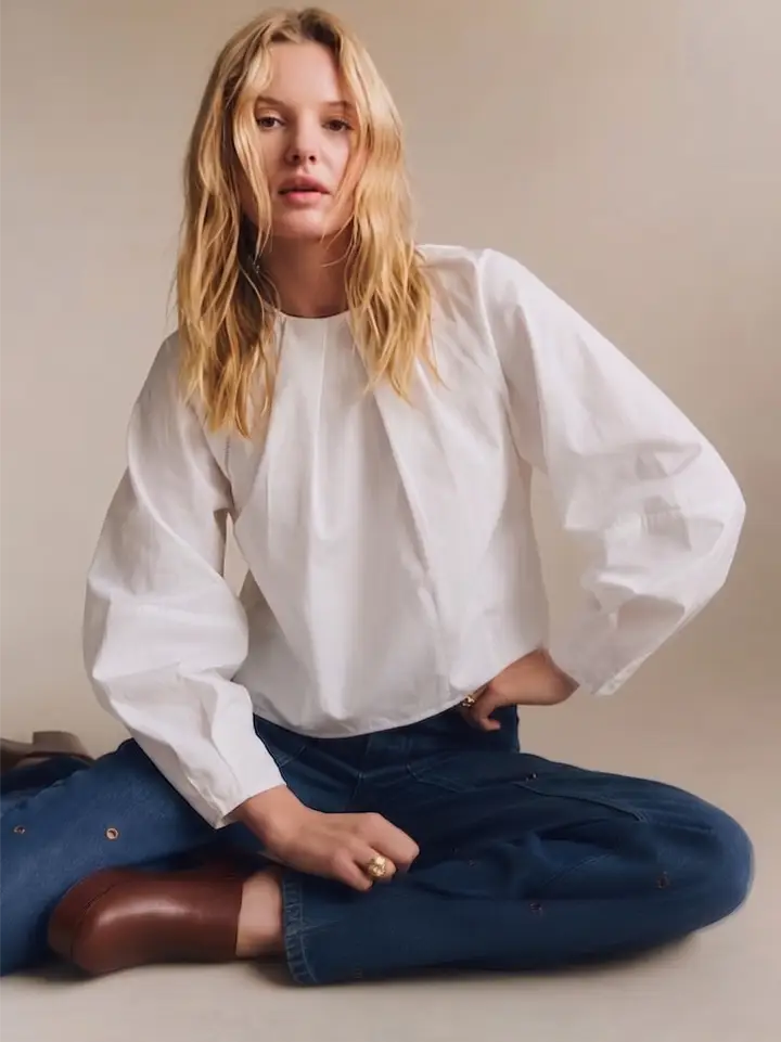 A woman sits cross-legged on a beige studio floor wearing a loose white long-sleeve blouse with voluminous sleeves, dark blue jeans, and brown ankle boots.