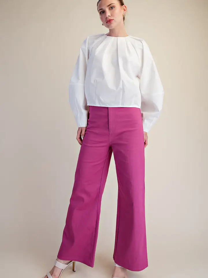 A model wearing a white long-sleeve blouse with dramatic puffed shoulders and bright magenta wide-leg trousers, standing against a plain beige studio background.