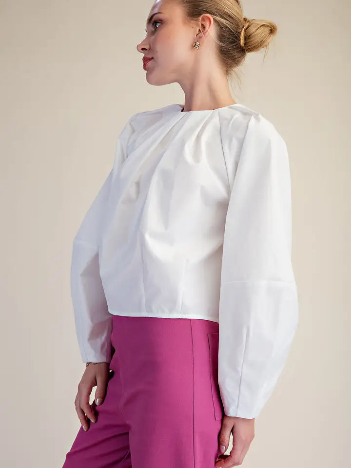 A woman wearing a white long-sleeve blouse with dramatic gathered shoulders and bright pink trousers, photographed against a plain warm beige studio background.