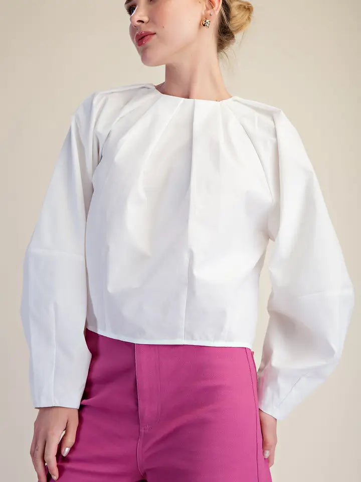 A woman wearing a white long-sleeve blouse with dramatic gathered shoulders and a high neckline, paired with bright pink pants, against a plain beige studio background.