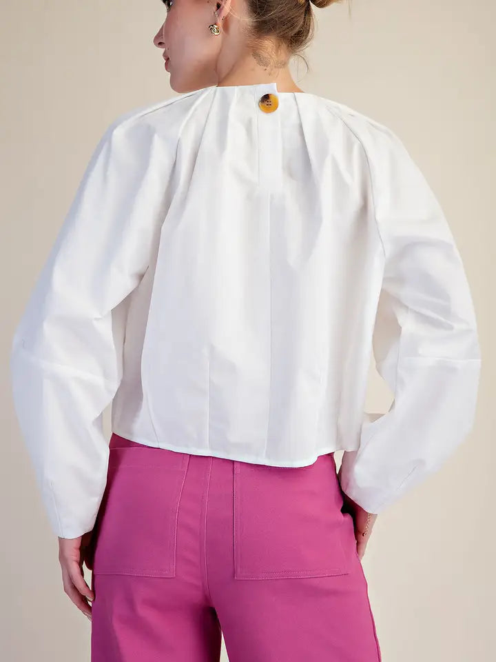 A woman shown from the back wearing a white long-sleeve blouse with a dramatic pleated neckline and a single decorative button, paired with bright pink trousers, against a plain beige studio background.
