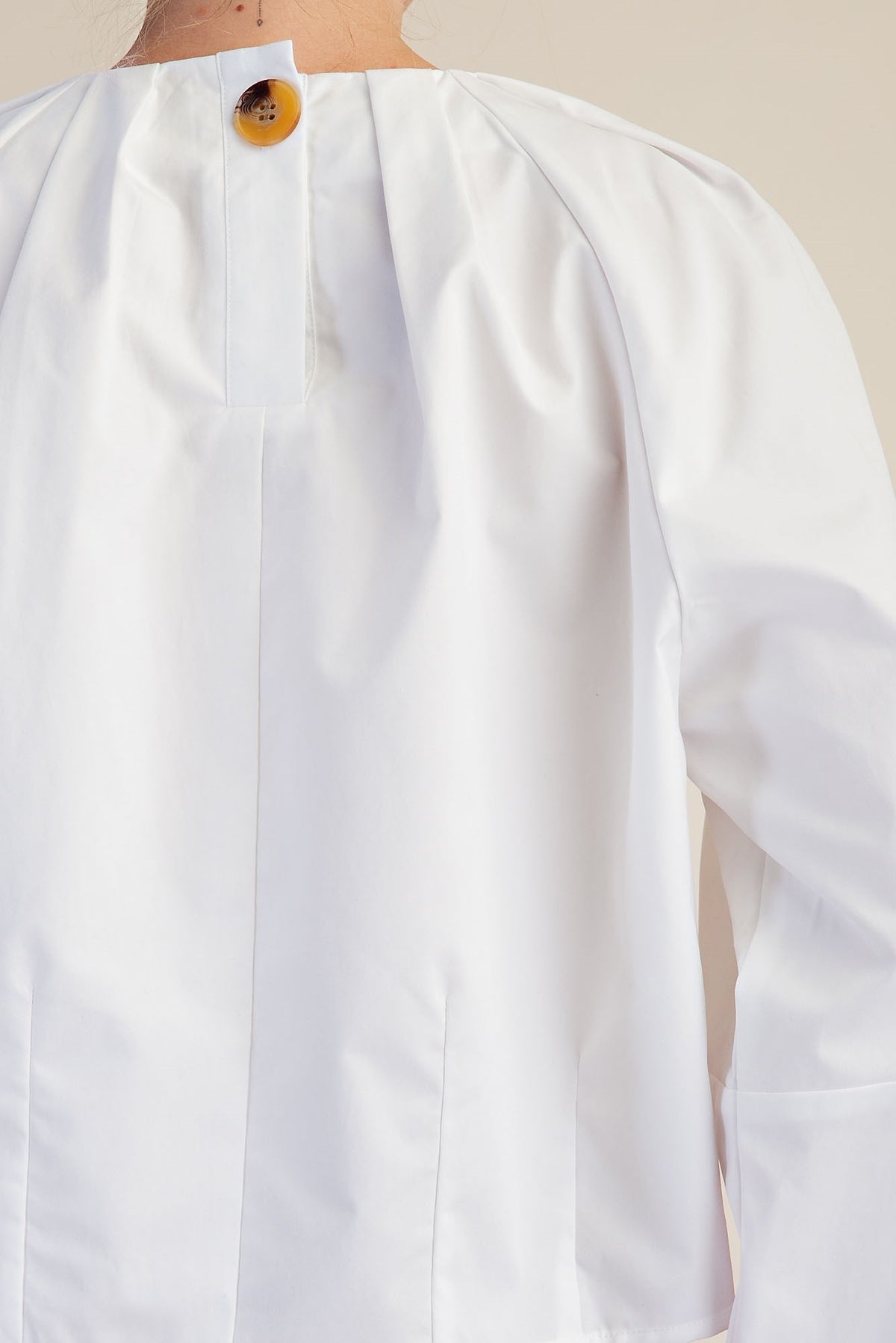 Close-up of the back of a white long-sleeve blouse with a gathered pleated neckline and a single round brown button, shown on a model against a light beige background.