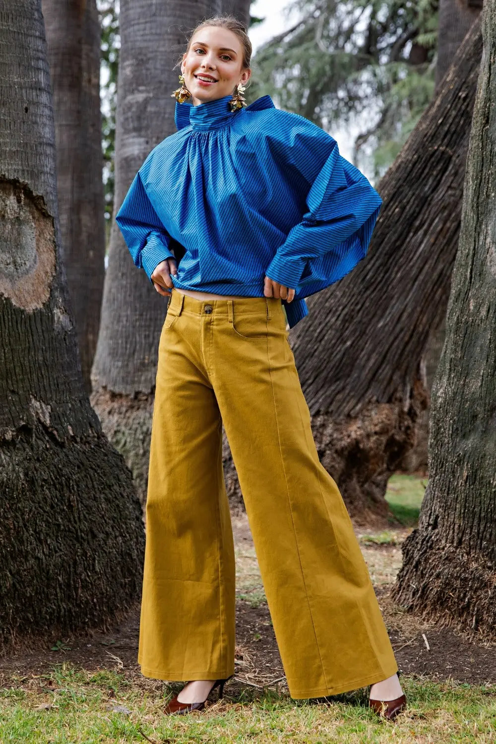 A woman stands outdoors among large tree trunks wearing a bright blue high-neck balloon-sleeve top and mustard yellow wide-leg pants.