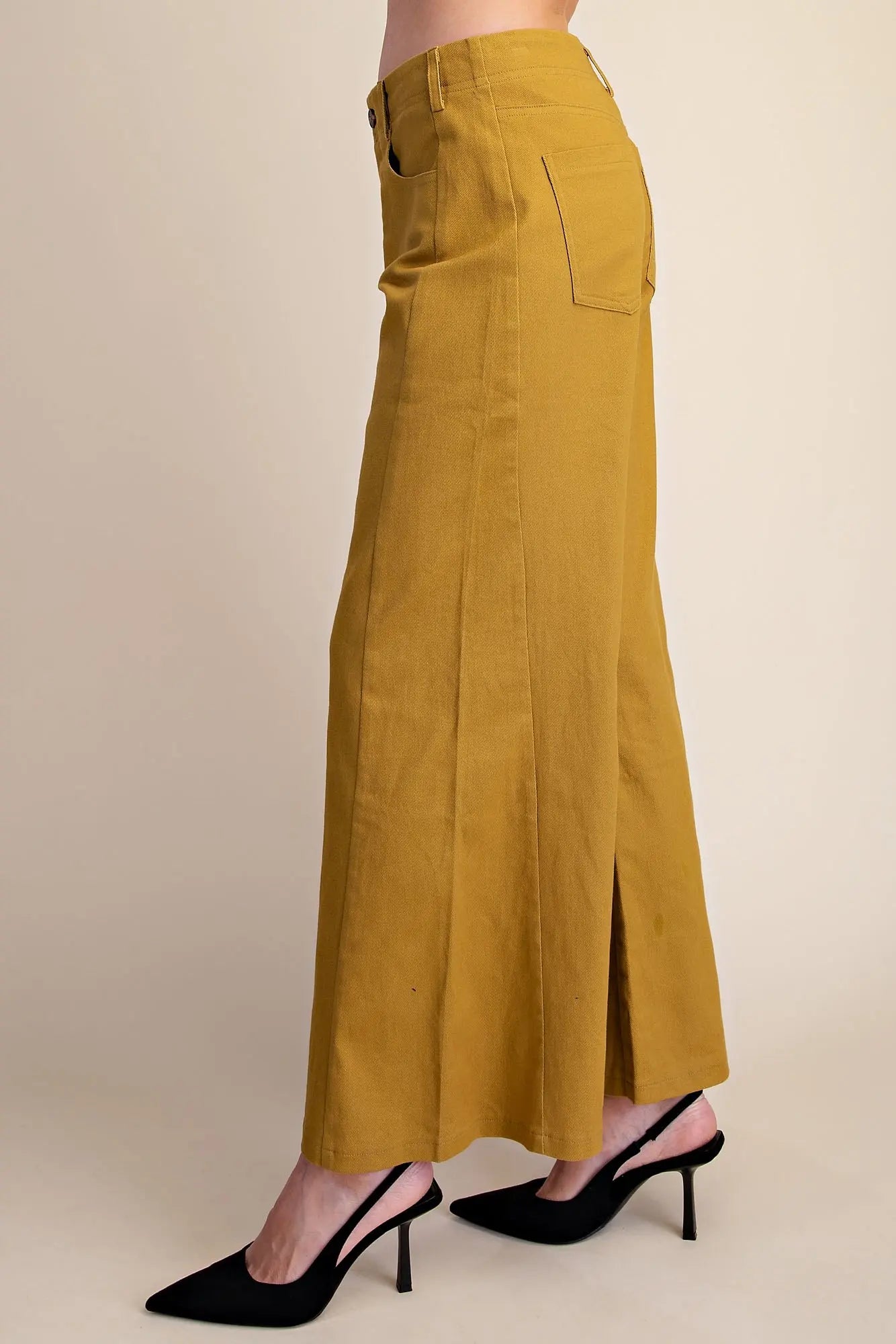 A person shown from the waist down wearing a mustard yellow wide-leg skirt with side slits and black pointed-toe slingback heels, against a plain beige studio background.