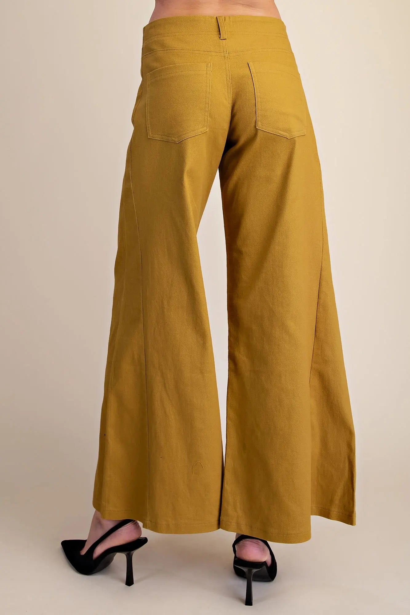 A person shown from the back wearing mustard yellow wide-leg pants and black slingback high heels against a plain beige studio background.