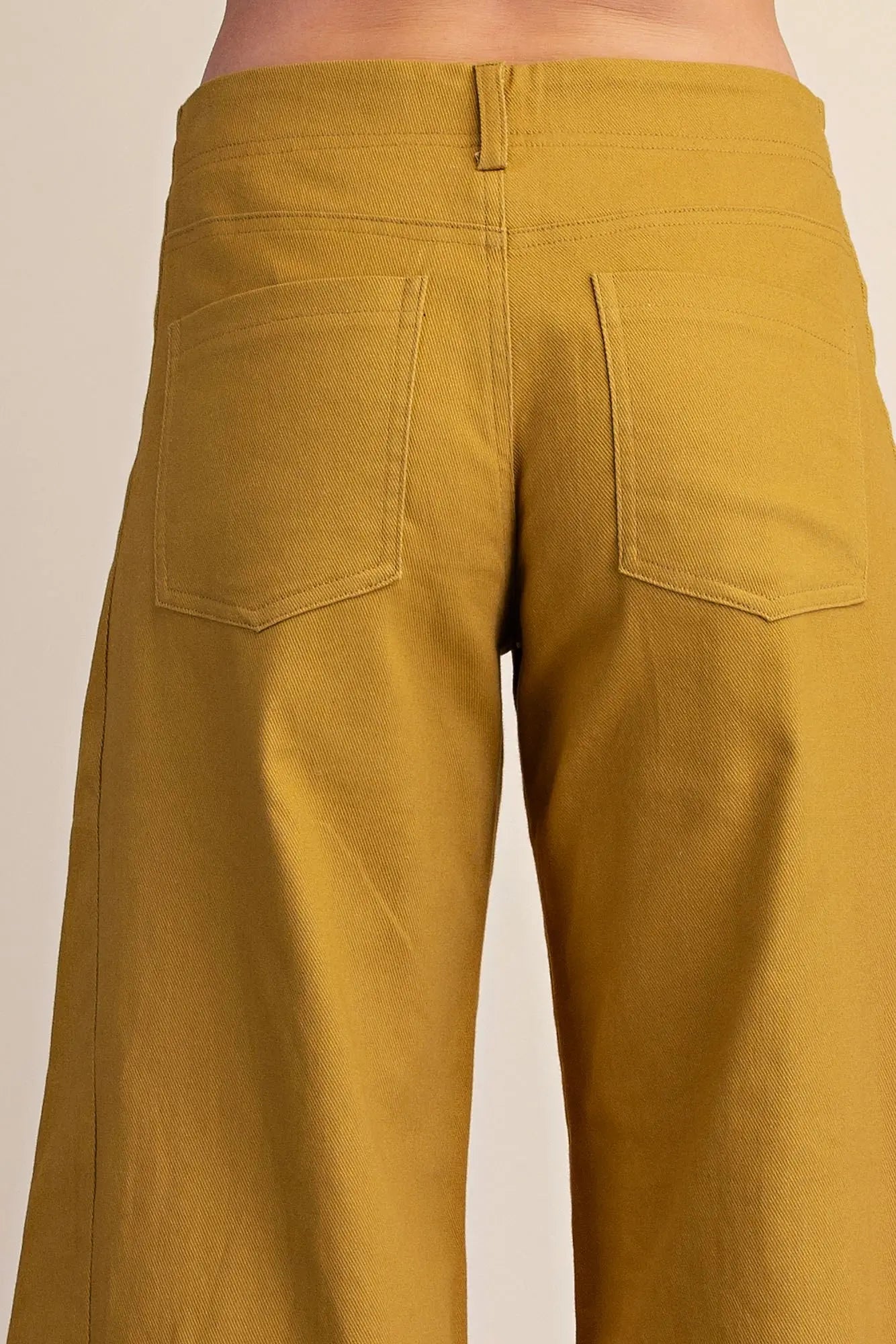 Close-up back view of a person wearing mustard yellow wide-leg pants, showing the waistband, button closure, and two back patch pockets against a plain light beige background.