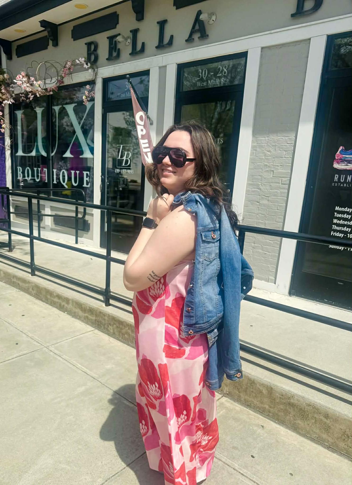 Woman in a floral dress and sunglasses standing in front of a store with 'Bella' and 'Lux Boutique' signs.