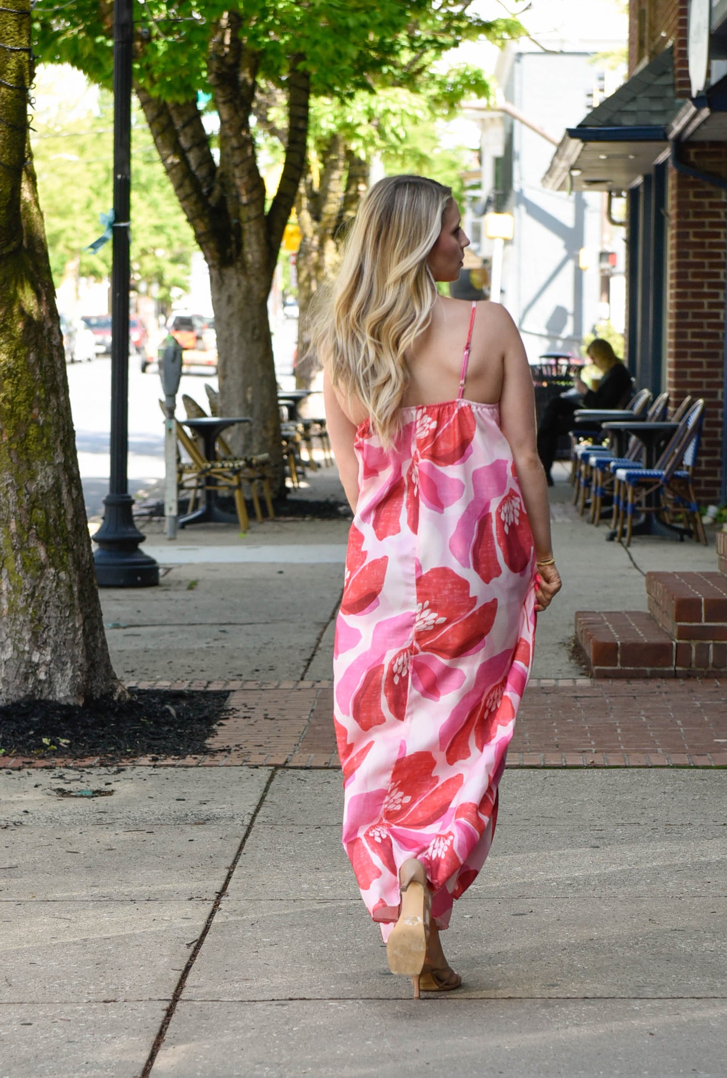 Woman in a pink floral dress walking on a sidewalk with trees and buildings in the background.