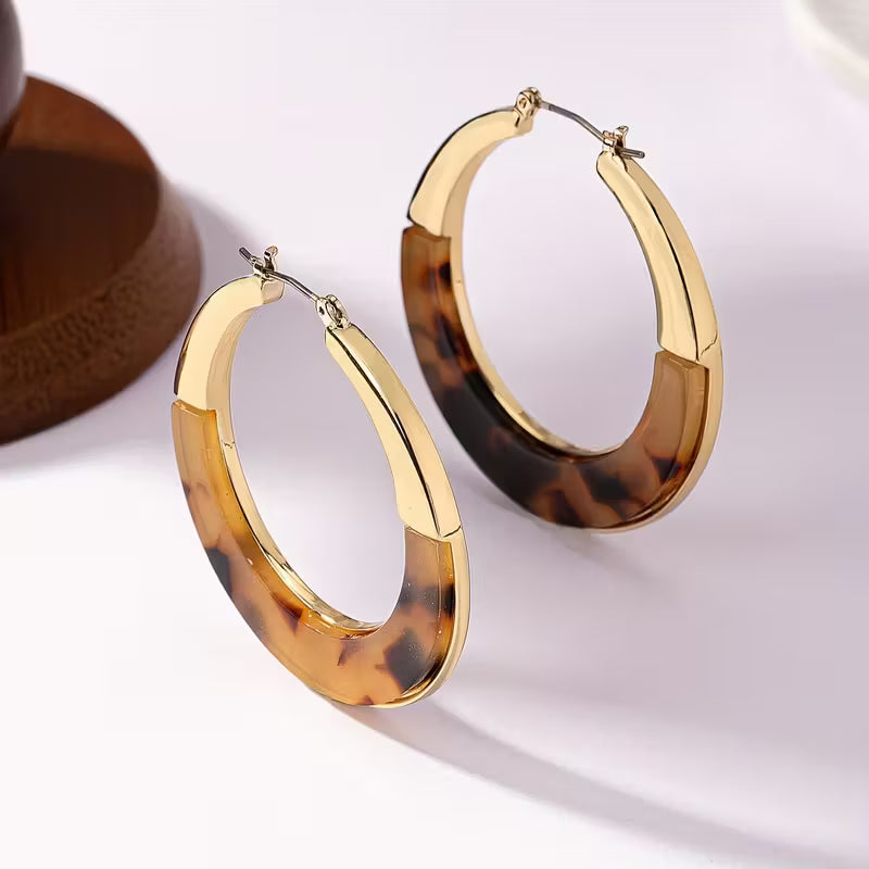 A pair of large hoop earrings with polished gold-toned metal and a tortoiseshell acetate inlay in amber, brown, and black, displayed on a light surface.