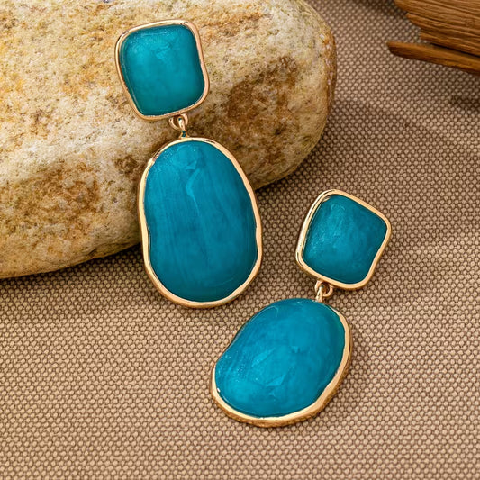 A pair of gold-tone drop earrings with large, organic-shaped turquoise blue stones and smaller square turquoise stones, displayed on a textured beige fabric next to a natural rock.