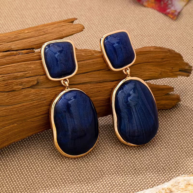 A pair of gold-tone drop earrings with deep blue stone inlays, displayed on a piece of natural wood over a textured beige fabric background.