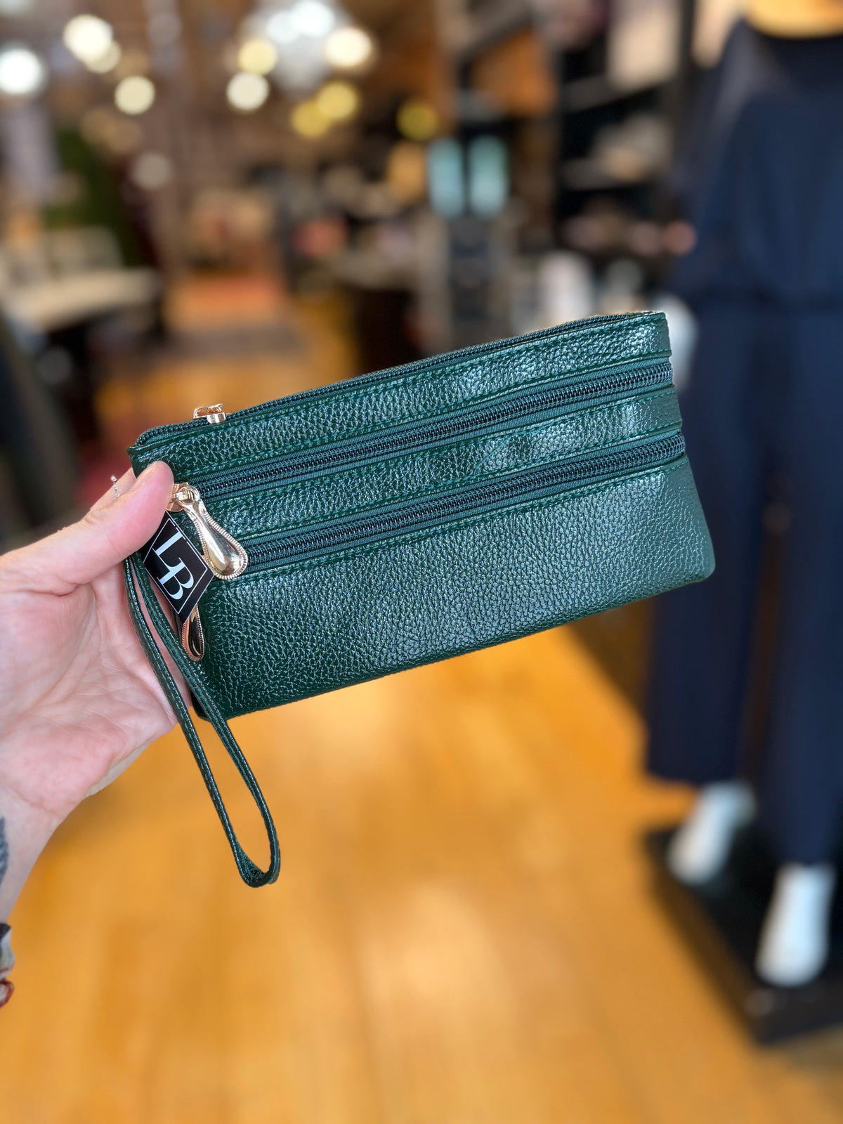 A dark green textured leather zip-around wristlet clutch with a wrist strap and silver-tone hardware, held by a hand in a blurred retail store setting.