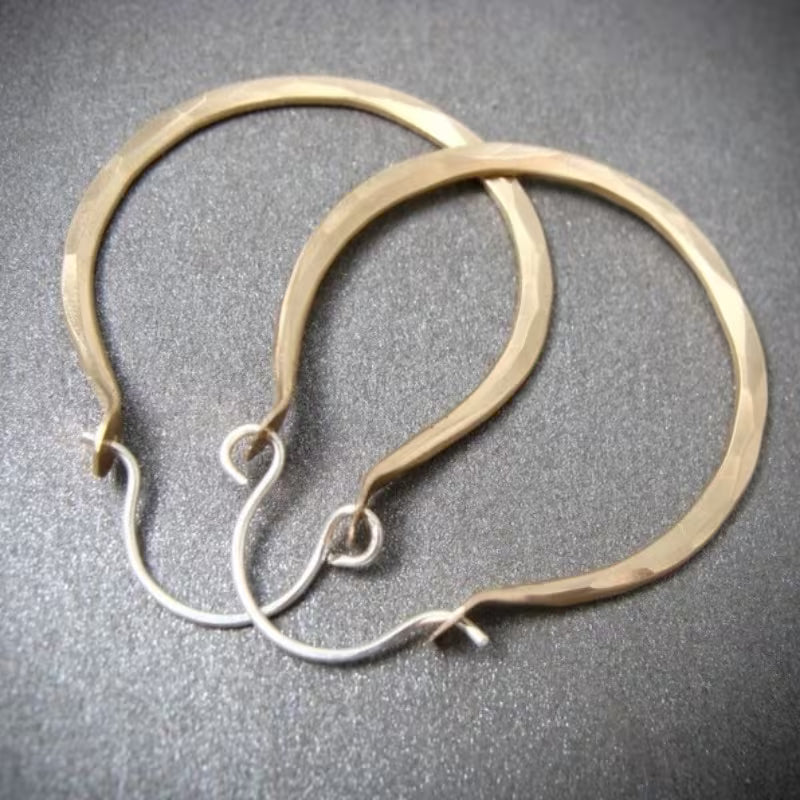 A pair of large hammered hoop earrings in a warm gold-tone finish, with silver-tone fishhook wires, displayed on a textured dark gray background.