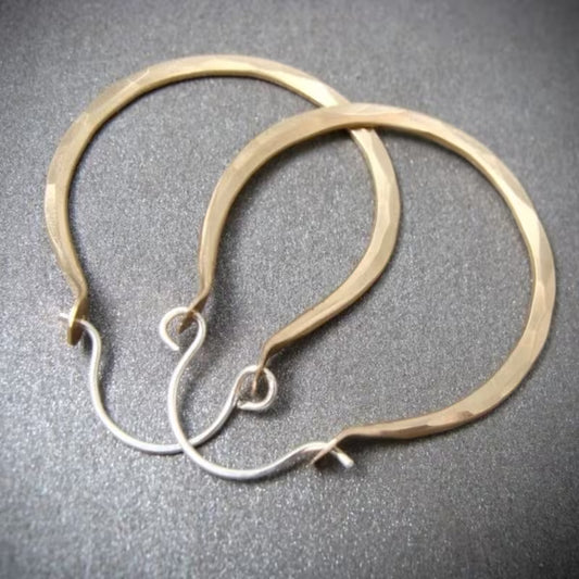 A pair of large hammered hoop earrings in a warm gold-tone finish, with silver-tone fishhook wires, displayed on a textured dark gray background.