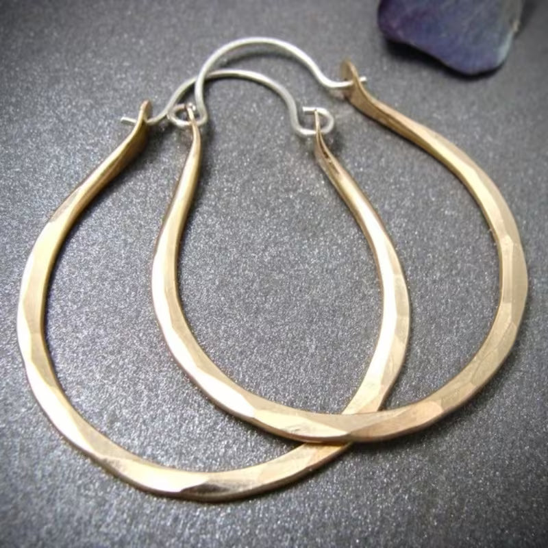 A pair of large hammered gold hoop earrings with silver fishhook backings, displayed on a textured gray surface.