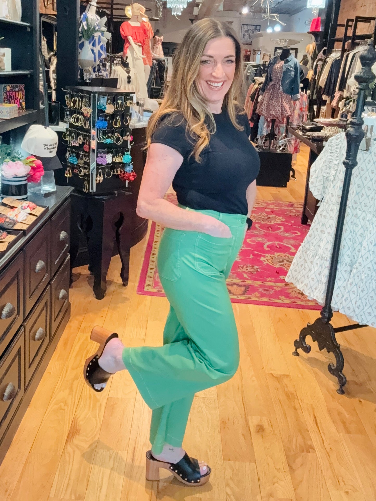 A woman stands in a boutique setting, wearing wide leg cropped pants in green with contrast stitching, paired with a black top.