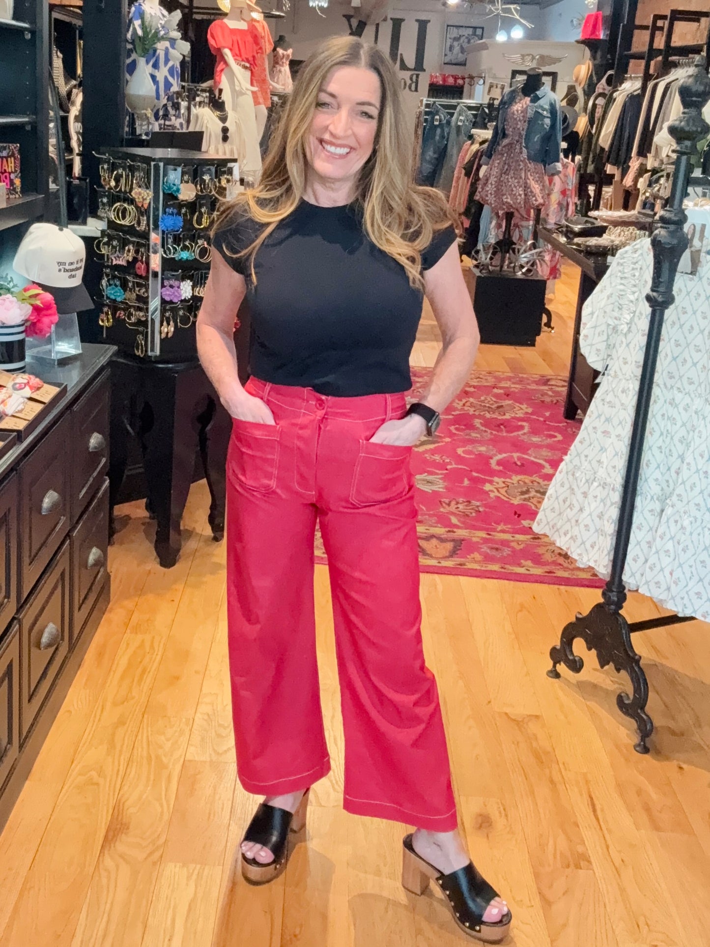 A woman stands in a boutique setting, wearing wide leg cropped pants in red with contrast stitching, paired with a black top.