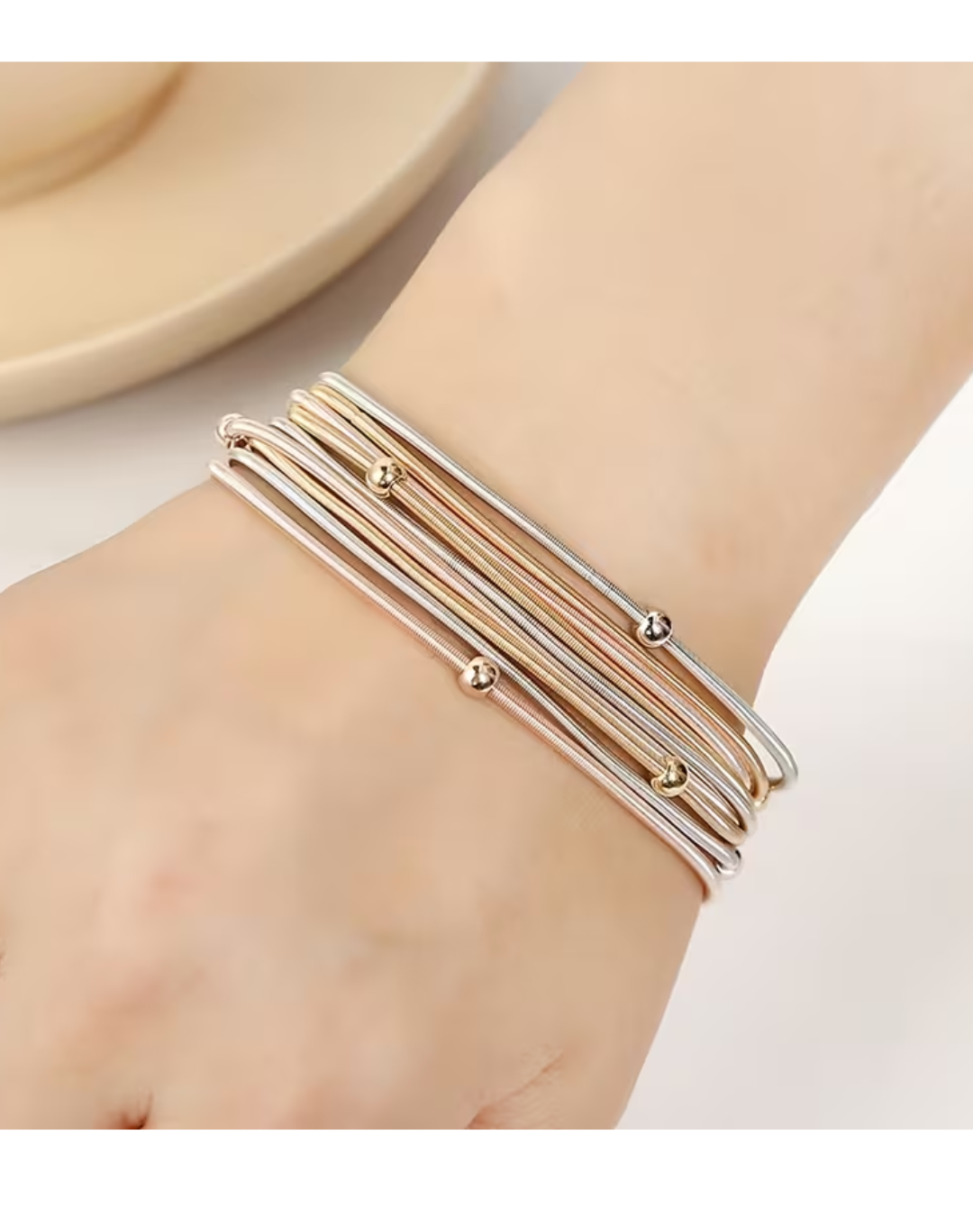A close-up of a person's wrist wearing five slim bangle bracelets in silver, gold, and rose gold tones, with small metallic bead accents.