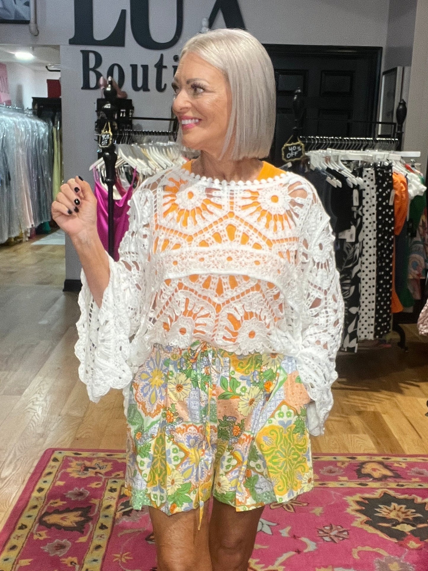A woman with a short blonde bob stands in a boutique wearing a white crochet poncho top with orange floral patterns and matching multicolor floral shorts. The background shows clothing racks and a store interior.