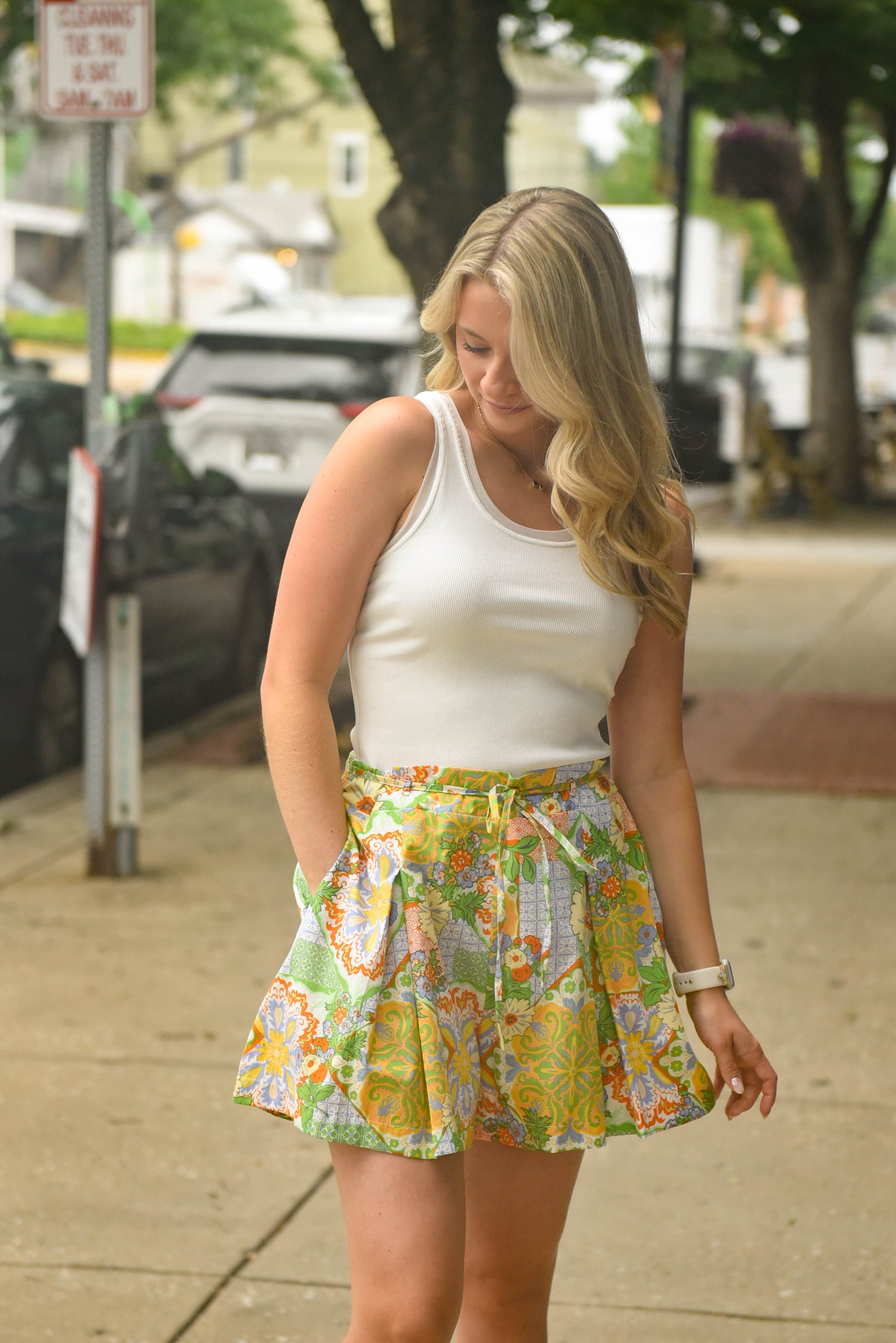 A woman standing on a city sidewalk wearing a white ribbed tank top and a colorful floral pleated mini skirt with a tie waist.