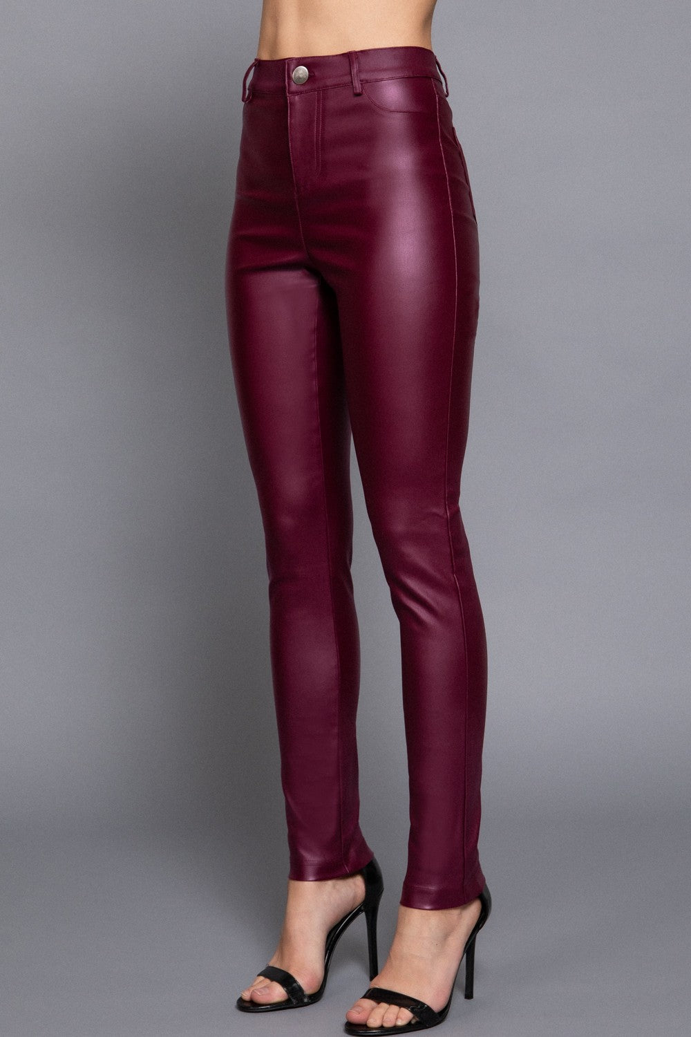 A model from the waist down wearing high-waisted, dark burgundy faux-leather skinny pants and black high-heeled sandals, standing against a solid gray studio background.